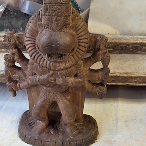 May include: A detailed wooden sculpture of a deity with a lion-like face and multiple arms. The figure is carved in a brown hue, with intricate details on the headpiece and body. The sculpture stands on a round base, showcasing a traditional artistic style.