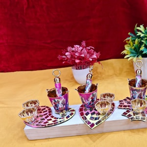 Op de afbeelding: Een set decoratieve serveerstukken, waaronder kleine emmers met sierlijke handvatten en bijpassende kleine kommen. De emmers en serveerbladen hebben een mozaïekontwerp met roze, rode en gouden accenten. Kunstplanten in witte potten staan op de achtergrond.