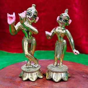 May include: Two gold-coloured statues of figures, possibly deities, stand on a wooden surface. The figures have detailed facial features, including painted eyes and pink accents on the ears and hands. They are posed in dynamic positions, suggesting movement.