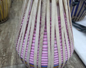 Könnte beinhalten: Eine rosa-beige Tabla-Trommel mit vertikalen beigefarbenen Lederriemen. Die Trommel hat eine zylindrische Form mit breiterem Boden und schmalerem Oberteil. Weitere Trommeln sind im Hintergrund zu sehen.