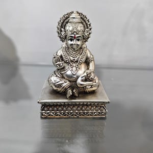 Könnte beinhalten: Eine silberfarbene Statue einer sitzenden Gottheit mit aufwendigem Schmuck und Heiligenschein. Die Figur steht auf einer dekorativen Plattform mit detaillierten Mustern. Die Statue hat einen ruhigen Ausdruck und einen roten Punkt auf der Stirn.