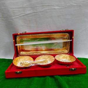 May include: A red velvet box containing a gold-coloured tray and three gold bowls. Each bowl features a decorative pattern and a small gold spoon. The set is designed for serving and is presented on a green surface.