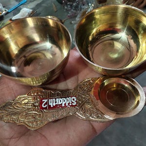 May include: Two polished brass bowls with horizontal ridged textures and a decorative brass spoon featuring an ornate handle with a red label that reads Siddarth 2. The items have a metallic gold finish and a smooth, reflective surface throughout.