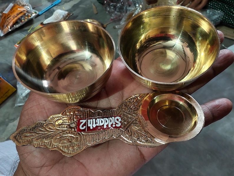 May include: Two polished brass bowls with horizontal ridged textures and a decorative brass spoon featuring an ornate handle with a red label that reads Siddarth 2. The items have a metallic gold finish and a smooth, reflective surface throughout.