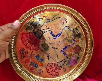 May include: A round, gold-colored metal tray with an intricate design of two birds and floral patterns. The birds are depicted in shades of blue, red, and orange, with the flowers in various colors. The tray has a decorative, ridged edge.