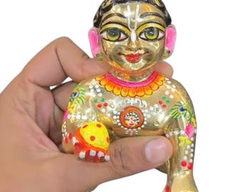 May include: A gold-colored figurine of a deity, held in a hand. The figurine has detailed facial features, including large eyes and a small smile. It is adorned with colorful paint and intricate patterns, with a small bun on top of the head.