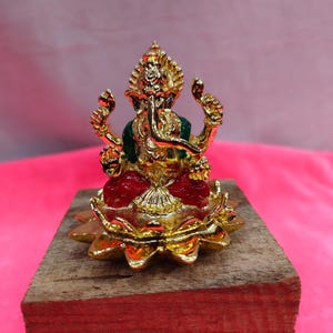 May include: A gold-colored statue of the Hindu deity Ganesha, seated on a red lotus flower. The statue has a green shawl and is mounted on a wooden block. The background is a bright pink.
