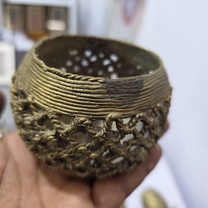 Pode incluir: Uma pequena tigela de latão antigo com um design vazado texturizado. A tigela tem uma forma arredondada com uma série de cristas horizontais perto do topo e um padrão vazado decorativo ao redor da base. A tigela é segurada em uma mão.