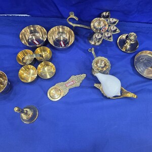 May include: A collection of brass items including two small bowls, a multi-tiered oil lamp, a bell, a small plate, a spoon, a decorative handle, and a white conch shell resting on a brass stand. The items are arranged on a blue fabric surface.
