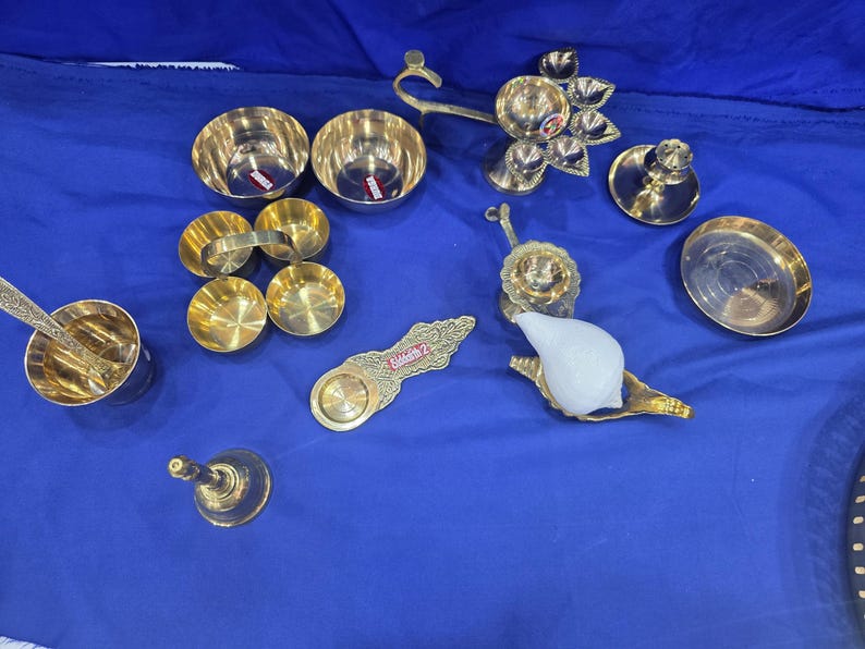 May include: A collection of brass items including two small bowls, a multi-tiered oil lamp, a bell, a small plate, a spoon, a decorative handle, and a white conch shell resting on a brass stand. The items are arranged on a blue fabric surface.