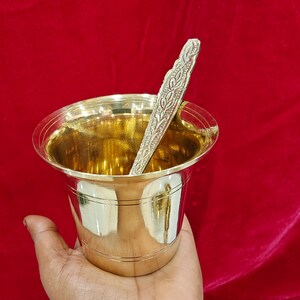May include: A polished brass cup with a wide, flared rim and horizontal line detailing around the body. Inside the cup rests a slender, textured brass spoon featuring an intricate, embossed leaf-like pattern along its entire handle length.