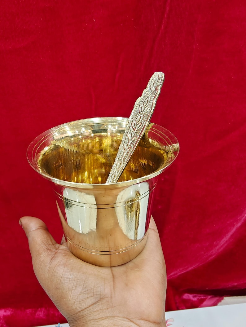 May include: A polished brass cup with a wide, flared rim and horizontal line detailing around the body. Inside the cup rests a slender, textured brass spoon featuring an intricate, embossed leaf-like pattern along its entire handle length.