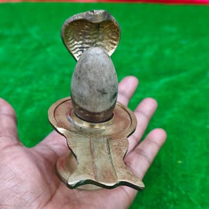 Puede incluir: Una escultura religiosa de latón con una capucha de cobra sobre una piedra ovalada, colocada sobre una base circular. La escultura se sostiene en una mano, con un fondo rojo y verde.