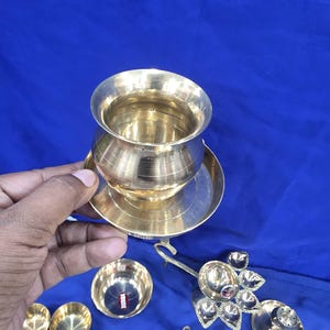 May include: A polished brass pot with a wide rim sits on a matching circular brass plate. Additional brass items include a small bowl with a red label, a multi-petaled oil lamp, and other small brass vessels arranged on a blue surface.