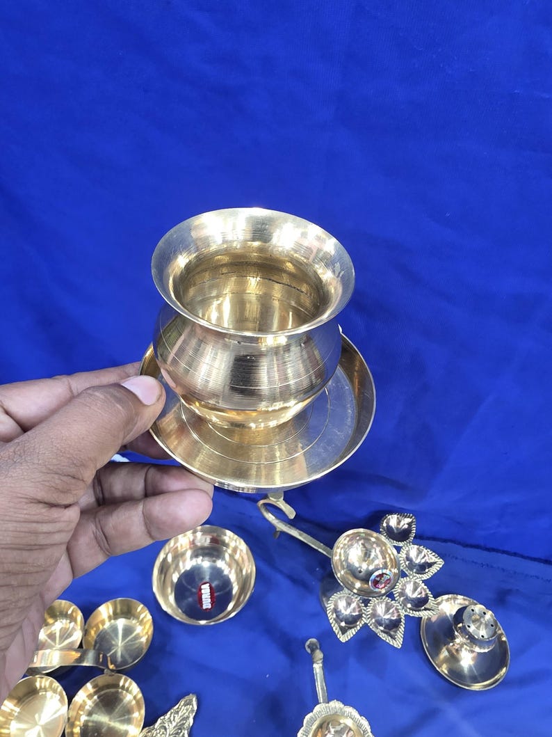 May include: A polished brass pot with a wide rim sits on a matching circular brass plate. Additional brass items include a small bowl with a red label, a multi-petaled oil lamp, and other small brass vessels arranged on a blue surface.
