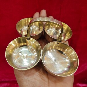 May include: A set of four small, round, polished brass bowls arranged in a square formation. The bowls are connected by a central, arched brass handle that spans across the middle, allowing for easy transport of the individual containers.