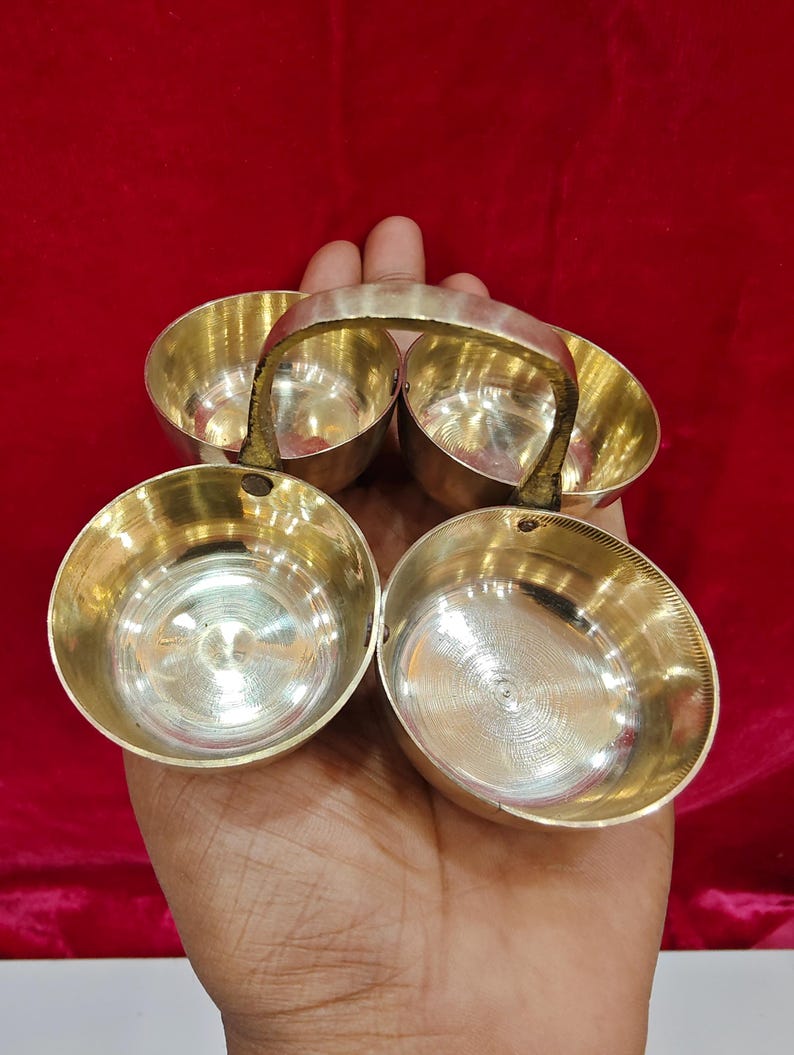 May include: A set of four small, round, polished brass bowls arranged in a square formation. The bowls are connected by a central, arched brass handle that spans across the middle, allowing for easy transport of the individual containers.