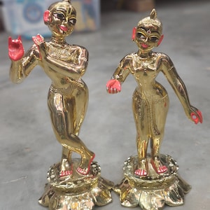 May include: Two gold-coloured statues of figures with red accents on their hands and feet. The statues are on ornate bases and have detailed facial features. The figures are posed gracefully, suggesting a religious or cultural theme.