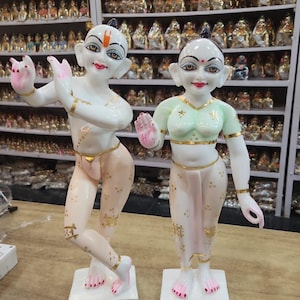 Puede incluir: Dos estatuas blancas y rosas de figuras con detalles dorados. Una figura está en pose de baile, la otra está de pie. Ambas tienen rasgos faciales pintados y están sobre bases cuadradas blancas.