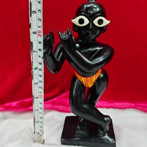 Pu&ograve; includere: Statuetta nera di una divinit&agrave; con occhi bianchi e perizoma giallo e arancione. La statuetta &egrave; su una base nera ed &egrave; alta circa 10 cm. Lo sfondo &egrave; rosso.