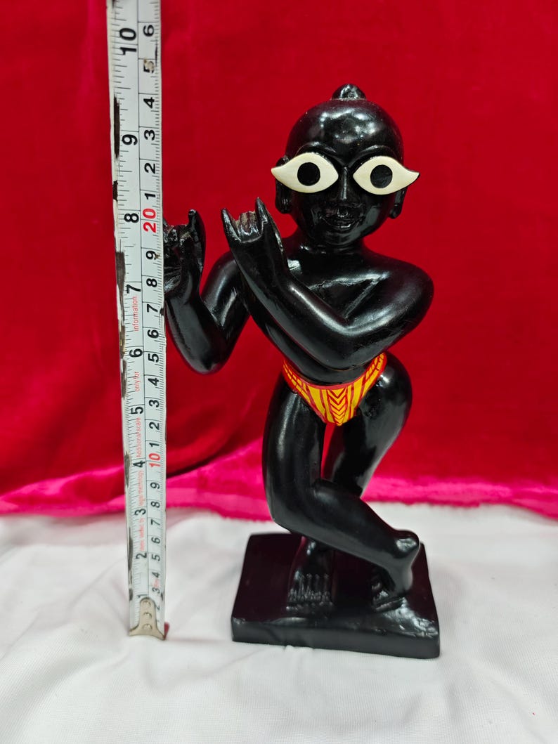 Pu&ograve; includere: Statuetta nera di una divinit&agrave; con occhi bianchi e perizoma giallo e arancione. La statuetta &egrave; su una base nera ed &egrave; alta circa 10 cm. Lo sfondo &egrave; rosso.