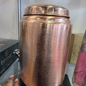 Puede incluir: Un recipiente cilíndrico de color cobre con tapa. El recipiente tiene un patrón floral en relieve alrededor del cuerpo. El recipiente está sobre una superficie negra.