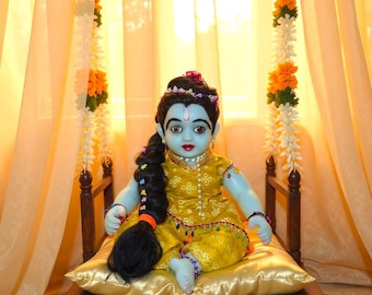 May include: A blue-skinned doll of a deity sits on a wooden swing with a gold satin pillow. The doll wears a yellow outfit with gold embellishments and a long black braid. The background includes floral garlands and sheer curtains.