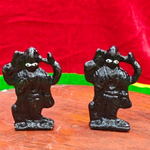 Puede incluir: Dos pequeñas figuras negras con ojos blancos y brazos levantados. Las figuras tienen una superficie texturizada y están sobre una superficie de madera. El fondo es de color rojo liso.