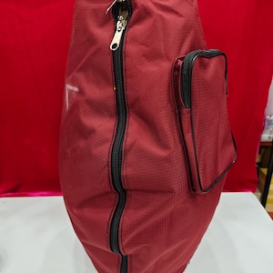 May include: A maroon bag with a black zip down the front and a smaller pocket with a black zip on the side. The bag has a textured surface and a black handle at the top.