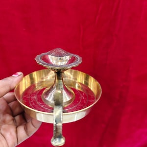 May include: A brass oil lamp with a handle, featuring a central bowl and a smaller, decorative upper bowl. The lamp has a polished finish and sits against a red background. The design is traditional and ornate.