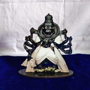 May include: A black and white statue of a deity with multiple arms, wearing a crown and white clothing. The statue has a fierce expression with red eyes and is standing on a round, dark base. The background is a blue surface.