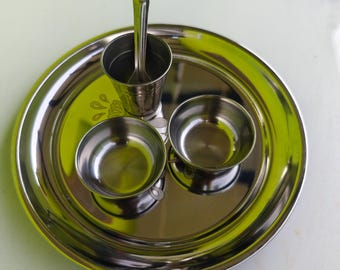 May include: A stainless steel thali set with a large round plate, two small bowls, a small cup, and a spoon. The set is reflective and has a simple, functional design. The thali is a traditional Indian serving plate.