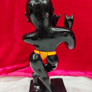 Pu&ograve; includere: Una statuetta nera di una persona in ginocchio, con un indumento giallo e rosso intorno alla vita. La statuetta &egrave; su una base rettangolare nera, con uno sfondo rosso.