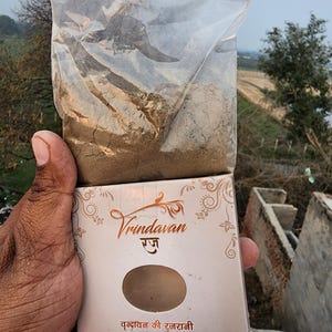 May include: A clear plastic bag filled with a brown powder, held open over a white box. The box has the word "Vrindavan" in a decorative script, with text below in a foreign language. The background shows a field and a cloudy sky.