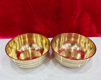 May include: Two small, gold-colored bowls with a red interior and a polished finish. The bowls have a rounded shape and are placed side by side. The bowls are likely used for serving or display.