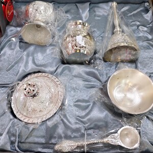 May include: A silver-colored decorative set, including a bell, small bowl, spoon, and other ornate pieces, all packaged in clear plastic within a gray satin-lined box. The items have detailed floral patterns.