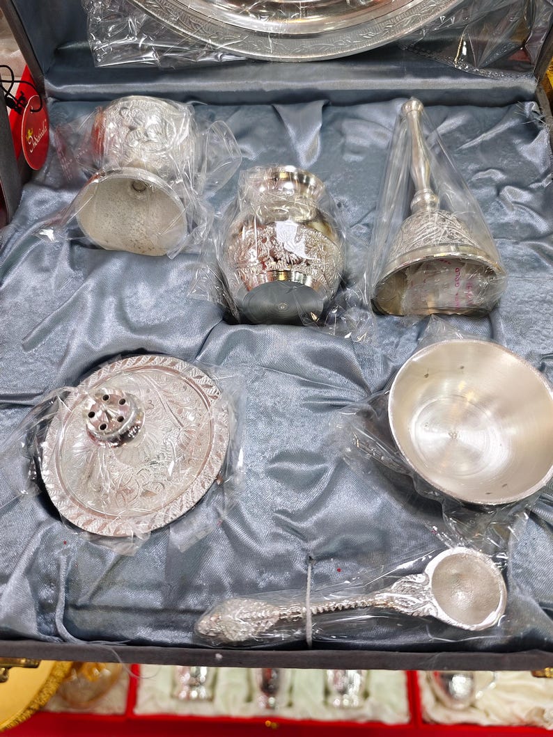 May include: A silver-colored decorative set, including a bell, small bowl, spoon, and other ornate pieces, all packaged in clear plastic within a gray satin-lined box. The items have detailed floral patterns.