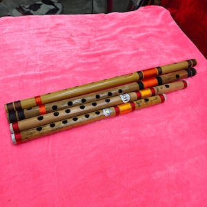 Puede incluir: Varias flautas de bambú con agujeros negros para los dedos y acentos coloridos en naranja, rojo y amarillo se disponen sobre una superficie rosa. Las flautas varían en tamaño y tienen bandas decorativas.