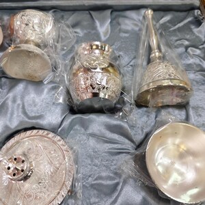 May include: A collection of ornate silver items, including a small vase, a bell with a handle, a small bowl, and a decorative plate. The items are packaged in clear plastic and rest on a gray satin-like fabric.