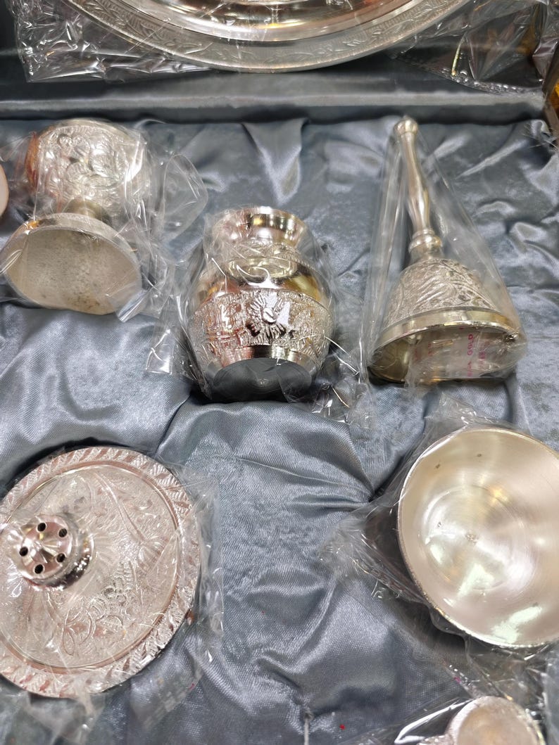 May include: A collection of ornate silver items, including a small vase, a bell with a handle, a small bowl, and a decorative plate. The items are packaged in clear plastic and rest on a gray satin-like fabric.