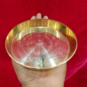 May include: A round, brass-coloured metal plate with a shallow, circular design. The plate is held in a hand against a red background. The plate has a shiny, reflective surface.