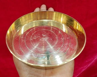 May include: A round, brass-coloured metal plate with a shallow, circular design. The plate is held in a hand against a red background. The plate has a shiny, reflective surface.