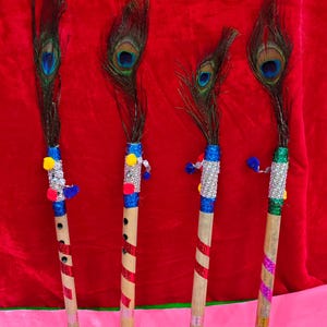 May include: Four decorative flutes with peacock feather accents. The flutes are made of bamboo, with red and white striped accents. The tops are adorned with blue, silver, and yellow embellishments. The background is a solid red.