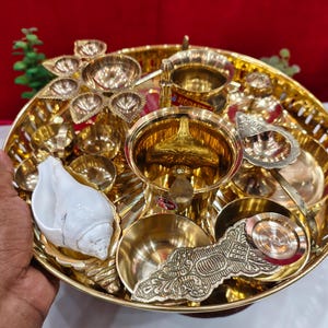 Puede incluir: Un thali de puja de metal dorado, o plato de ofrendas, lleno de varios artículos religiosos. El thali incluye pequeños cuencos, una concha y elementos decorativos. Los artículos están dispuestos en una bandeja redonda con un borde detallado.