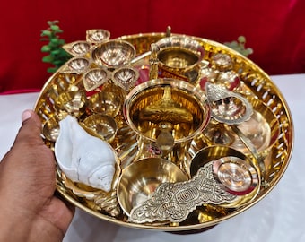 May include: A gold-colored metal puja thali, or offering plate, filled with various religious items. The thali includes small bowls, a conch shell, and decorative elements. The items are arranged on a round tray with a detailed edge.