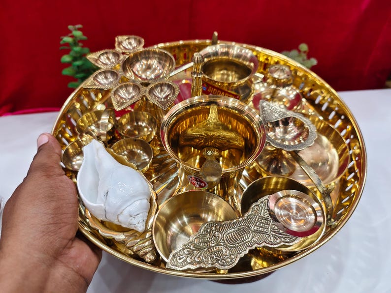May include: A round brass tray containing a collection of brass items including a central bowl, several small bowls, a multi-wick lamp, a decorative handle, and a white conch shell. The tray features a perforated rim and a polished gold-tone finish.
