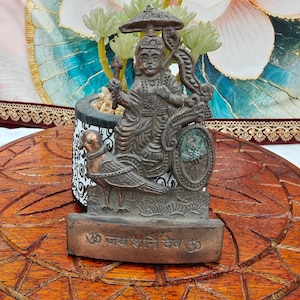 Op de afbeelding: Een bronzen beeld van een godheid op een door een vogel getrokken strijdwagen. Het beeld heeft ingewikkelde details en is gemonteerd op een rechthoekige basis met tekst. De achtergrond omvat een decoratieve pot met planten en een bloemmotief.