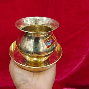 May include: A shiny, golden brass pot with a wide rim and a rounded body, sitting on a matching circular base. The pot has a sticker with text on it. The background is a solid red colour.
