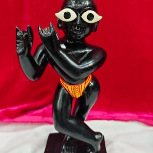 Pu&ograve; includere: Una statuetta nera di una divinit&agrave; con grandi occhi bianchi e un perizoma a motivi gialli e rossi. La statuetta &egrave; in posa in una posizione simile alla danza, con le braccia alzate e le dita ricurve. La base &egrave; di forma rettangolare nera.