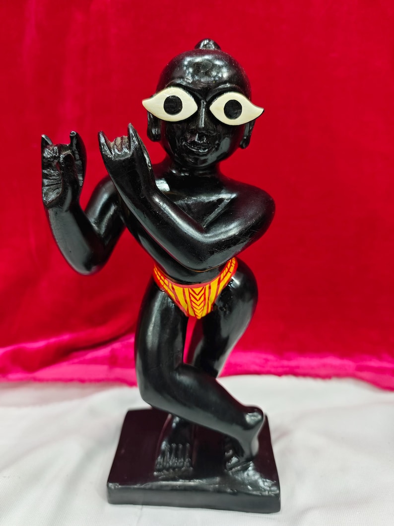 Pu&ograve; includere: Una statuetta nera di una divinit&agrave; con grandi occhi bianchi e un perizoma a motivi gialli e rossi. La statuetta &egrave; in posa in una posizione simile alla danza, con le braccia alzate e le dita ricurve. La base &egrave; di forma rettangolare nera.